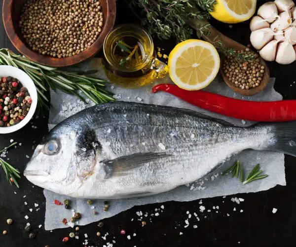 Poisson entier de dorade cru posé sur le plan de travail du restaurant la Table de 3 à Bordeaux, entouré de citron, piment rouge, herbes aromatiques, huile d’olive et épices.