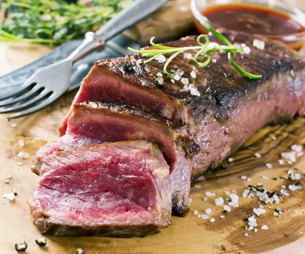 Tranches de bœuf saignantes au restaurant La Table de 3 de Bordeaux, parsemées de fleur de sel et de romarin, servies sur une planche en bois avec une sauce d'accompagnement et une fourchette.