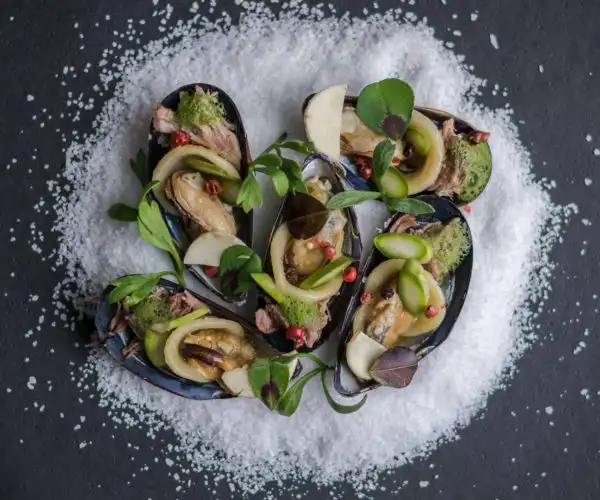 Cinq moules farcies délicatement dressées par notre équipe en cuisine de la Table de 3 à Bordeaux, avec des herbes fraîches, des baies roses et des jeunes pousses, présentées sur un dôme de gros sel blanc sur un fond sombre.