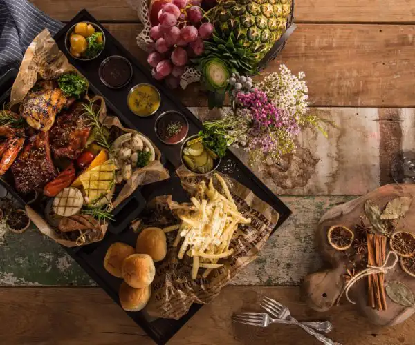 Un grand plateau de grillades mixtes proposé au restaurant La Table de 3 à Bordeaux, comprenant de la viande rôtie, des légumes grillés, des frites croustillantes et des petits pains, servi avec un assortiment de sauces et décoré de fruits frais sur une table en bois rustique.