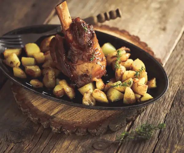 Une souris d'agneau braisée et laquée servie au restaurant bordelais La Table de Trois, servie dans un plat ovale noir avec des pommes de terre rissolées et du thym frais, le tout posé sur un billot de bois rustique.