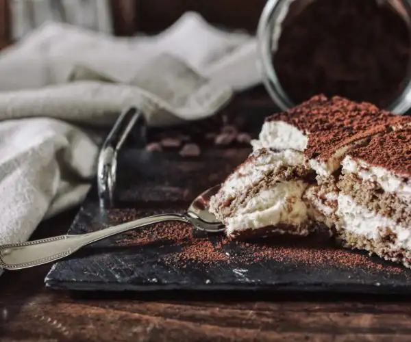 Une part généreuse de tiramisu traditionnel, recette secrète de notre cheffe à la Table de Trois de Bordeaux, saupoudrée de cacao amer, servie sur une ardoise noire avec une cuillère argentée, révélant les couches de crème mascarpone et de biscuits imbibés au café.